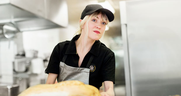 Blond kvinna i svarta arbetskläder och keps arbetar i ett storkök.