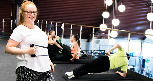 Fyra kollegor i ett gym. Kvinna som tittar i kameran och samtidigt drar i gummiband för att stärka armar och överkropp.